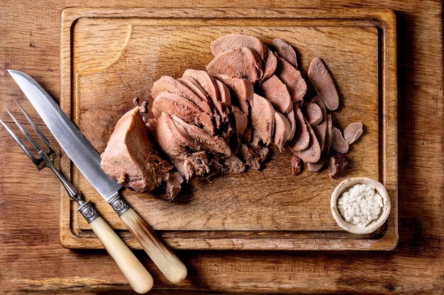 Baked beef tongue sliced, serving with horseradish sauce and meat knife on wooden cutting board over wood surface