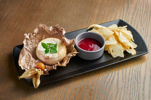 Baked Camembert on a black plate on a wooden surface with a croutons and jam.Close up view. Film effect during post. Soft focus