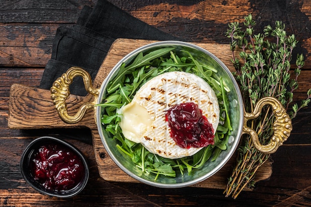 Baked Camembert Brie cheese with a cranberry sauce and garnished with arugula salad in a skillet Wooden background Top view