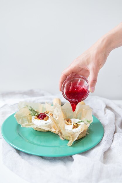 Baked camembert with walnuts, honey and rosemary wrapped in parchment lies on a green plate, a girl pours cranberry jam into the cheese, delicious and healthy food.