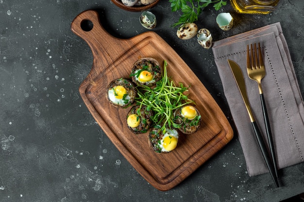 Baked champignons with quail eggs and microgreen garnish on a black plate top view