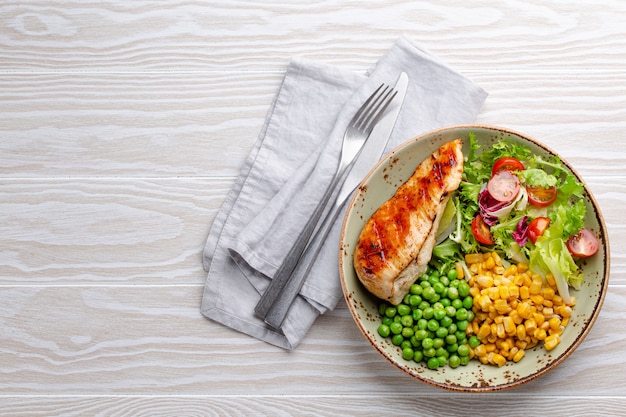 Baked chicken breast on plate with fresh salad, green peas and corn, white wooden background, top view. Healthy fitness meal with chicken fillet, balanced in proteins and carbs. Overhead, copy space