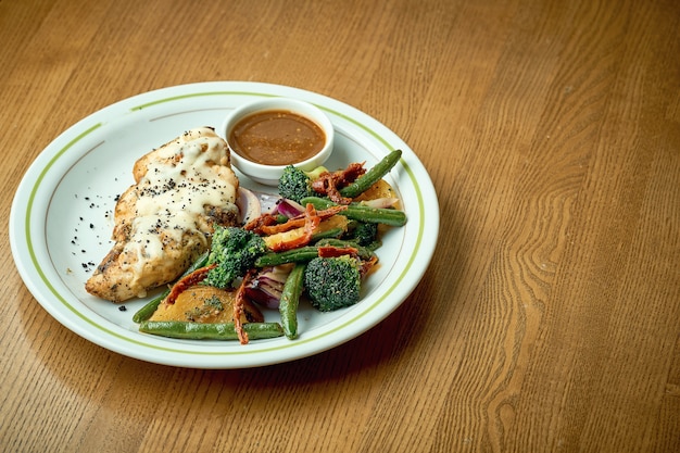 Baked chicken breast with cheese and boiled vegetables, broccoli in a white plate on a wooden table