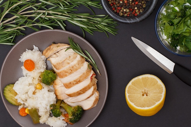 Baked chicken breasts or fillet on plate with rosemary, bowls with sauce and allspice berries, lemon and knife on black background. Top view.