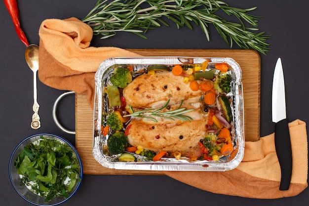 Запеченные куриные грудки или филе с овощами и зеленью в металлическом контейнере на деревянной разделочной доске. Стеклянная миска с соусом, ложкой и ножом. Вид сверху.