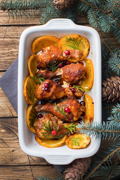 Photo baked chicken drumstick with oranges and cranberries in a baking sheet on a wooden background.