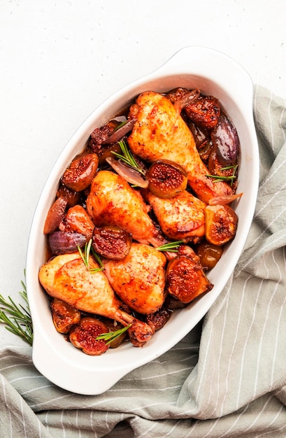 Baked chicken legs drumsticks with figs and red onions in ceramic baking dish white kitchen table background top view