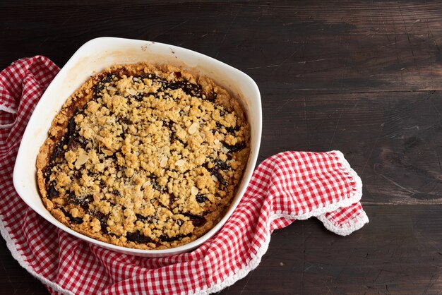 Baked crumble pie with blue plum in a white ceramic plate on a wooden table