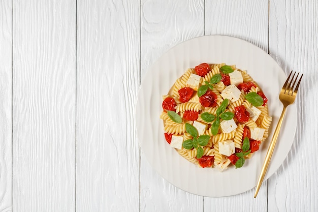 Baked feta cheese fusilli pasta with cherry tomatoes and basil on a white plate on a wooden table, flat lay, free space, italian cuisine