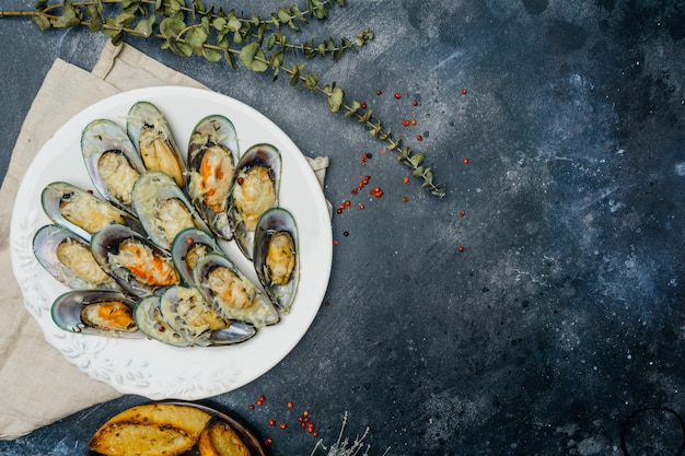 Baked green mussels with parmesan and garlic croutons on a white plate on a dark background. Copy space.