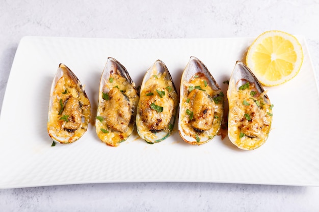 Baked Greenshell mussels in half a shell with cheese, parsley and lemon on a white plate. Traditional hot appetizer. Close-up, top view.