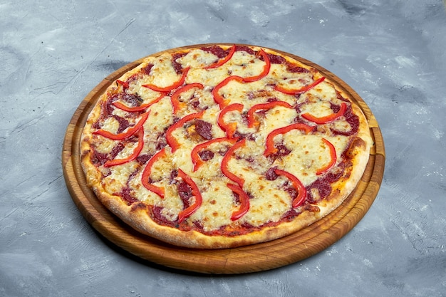 Baked italian pizza with smoked sausages, pickled cucumbers, salami and tomatoes on a wooden tray on a gray background.