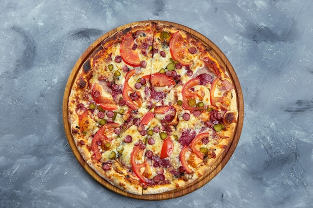 Baked italian pizza with smoked sausages, pickled cucumbers, salami and tomatoes on a wooden tray on a gray background.