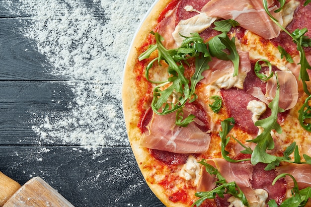 Baked pizza with salami, prosciutto and chicken with red sauce and melted cheese on a black wooden table in a composition with ingredients. Top view