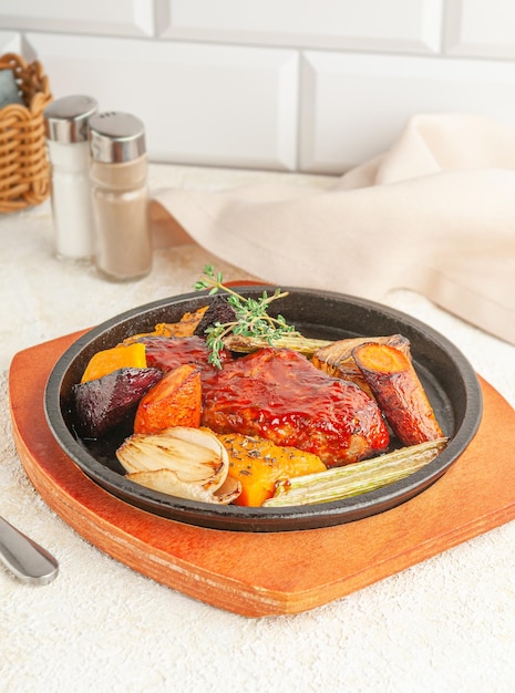 Baked pork in honey sauce. With grilled vegetables and thyme. Served in a frying pan. Nearby is a beige napkin and spices. Light background.
