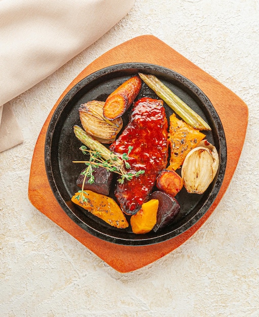 Baked pork in honey sauce. With grilled vegetables and thyme. Served in a frying pan. Nearby is a beige napkin. View from above. Light background