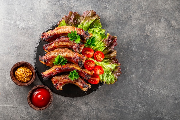 Baked pork ribs with vegetables mustard and ketchup on gray background top view free space for text