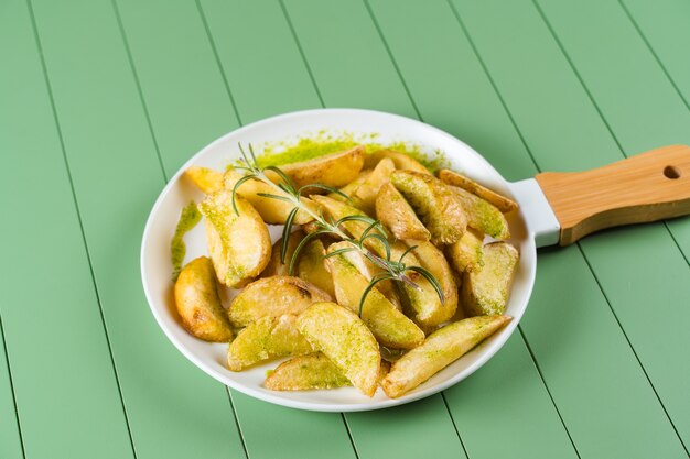 Baked potatoes with green sauce and rosemary on a white plate in the form of a frying pan. Fried potatoes with pesto sauce on a green table.