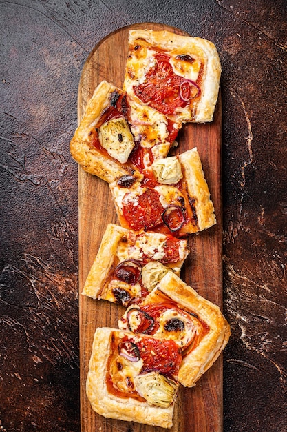 Foto pizza crostata di pasta sfoglia al forno con mozzarella di carciofi pomodori e formaggio affettato su una tavola di legno sfondo scuro vista dall'alto