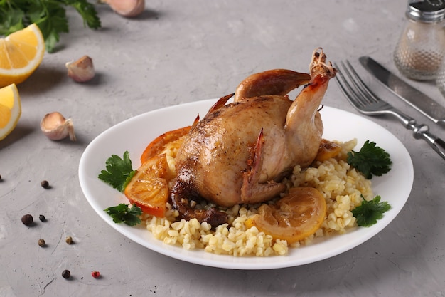 Baked quails with lemon and orange served with bulgur on a white plate on a gray background, Closeup.