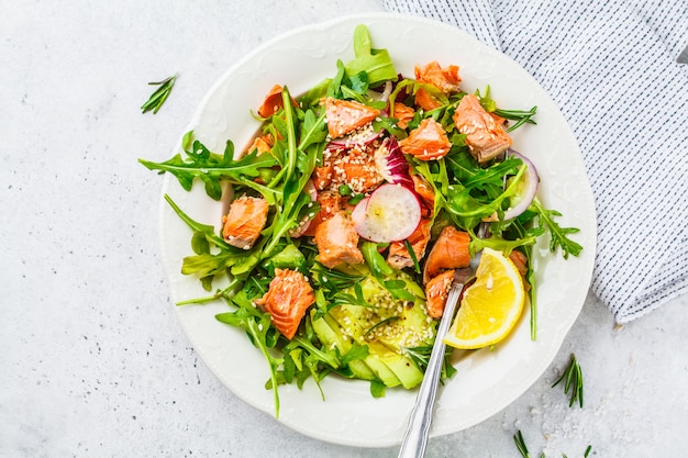 Baked red fish salad with avocado, cucumber, radish and arugula in white plate, top view.