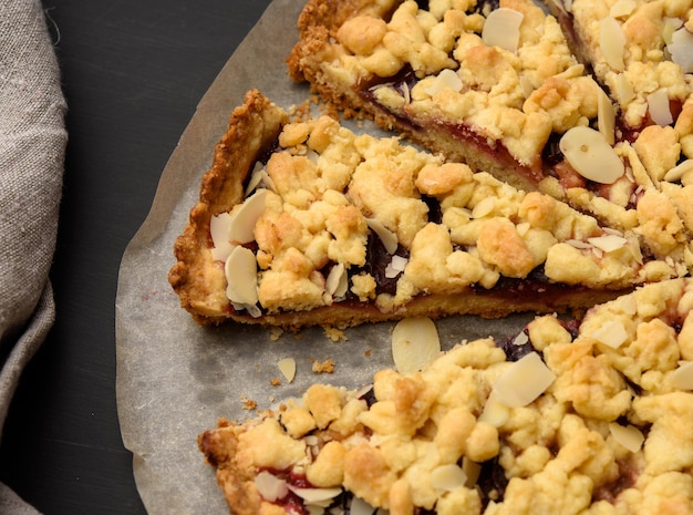 baked round crumble pie with plum cut into pieces on a black background close up
