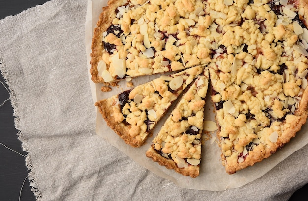 Baked round crumble pie with plum cut into pieces on a black background, top view