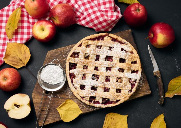 Печеный круглый традиционный яблочный пирог на коричневой деревянной доске и свежие красные яблоки сверху