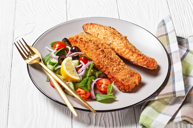 Baked salmon fillets breaded with Japanese panko crumbs served with vegetable salad and lemon wedge on a plate on a wooden table