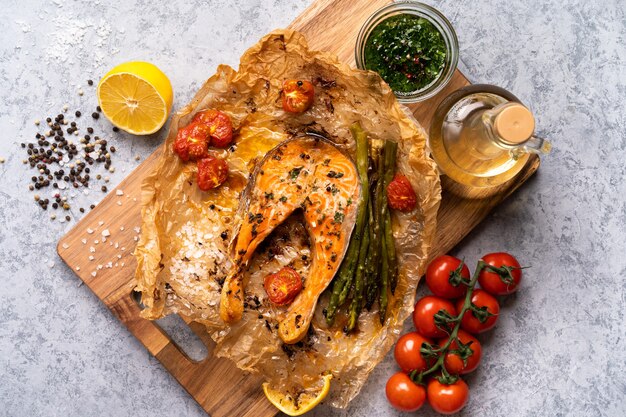 Trancio di salmone al forno su pergamena con verdure, asparagi, pomodori su un tavolo grigio.