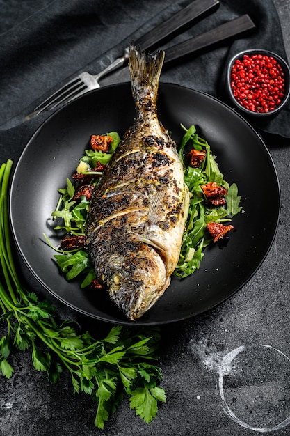 Baked sea bream fish with arugula salad and tomatoes. Black background. top view