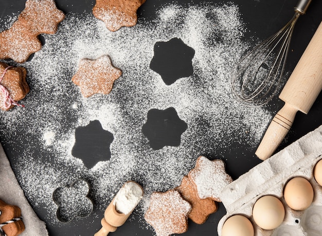 Запеченное имбирное печенье в форме звезды, посыпанное сахарной пудрой, на черном столе и ингредиенты, вид сверху