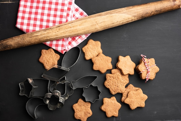 Baked star shaped gingerbread cookies, wooden rolling pin and metal cutters on a black table, top view