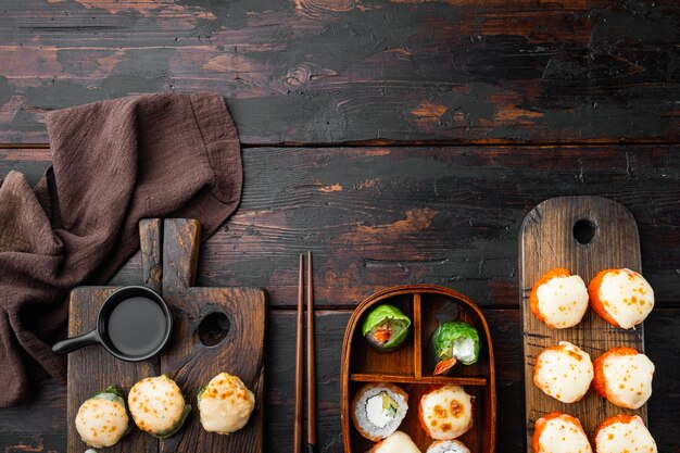 Rotoli di sushi maki al forno con salmone, granchio, cetriolo, avocado, uova di pesce volante e set di salsa piccante, su vecchio sfondo di tavolo in legno scuro, vista dall'alto piatta, con copyspace e spazio per il testo