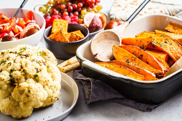 Patate dolci al forno, cavolfiore, frutta, insalata di verdure e tortilla con verdure su uno sfondo bianco.