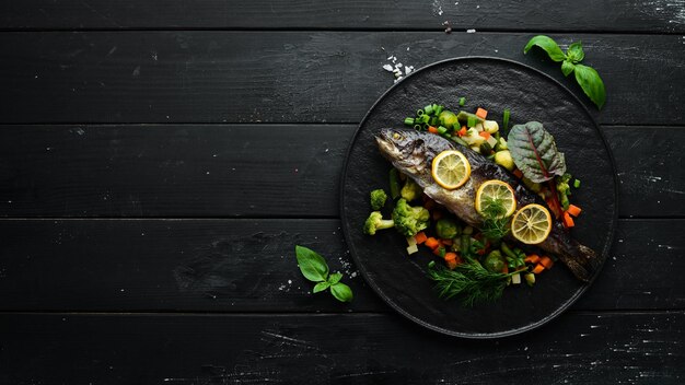 Baked trout fish with vegetables and lemon on a black plate Top view Free copy space