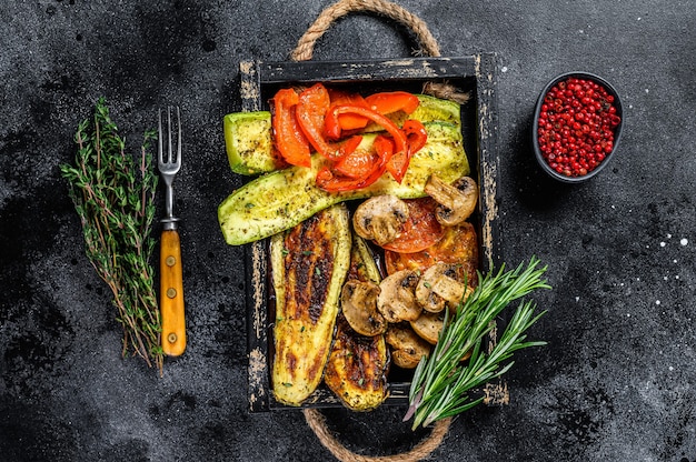 Baked vegetables bell pepper, zucchini, eggplant and tomato  in a wooden tray. Black wooden background. Top view.