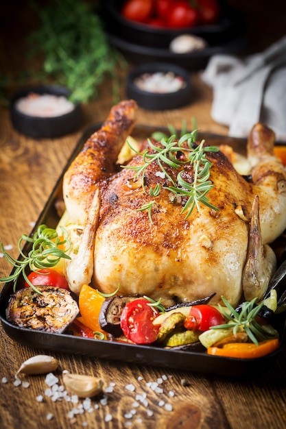 Baked whole chicken with vegetables and herbs close-up on a plate on a table.