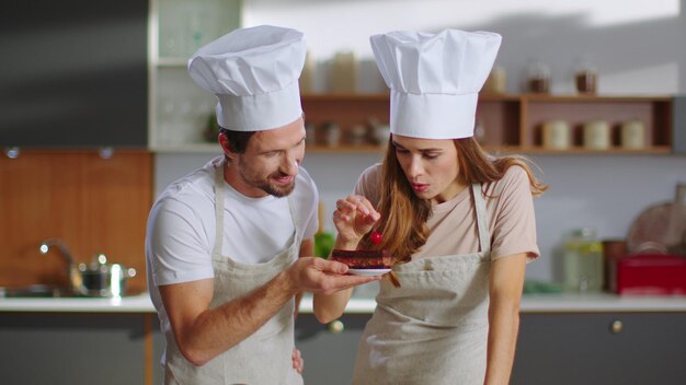 Baker and assistant putting cherry on cake at kitchen Couple of confectioners