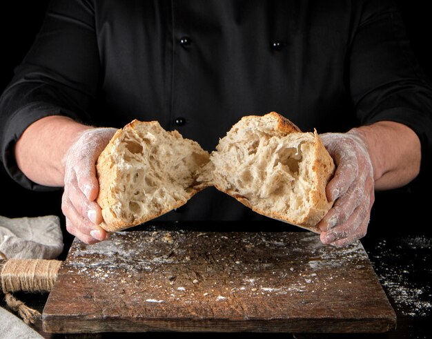 baker in black uniform broke in half a whole baked loaf of white wheat flour bread close up