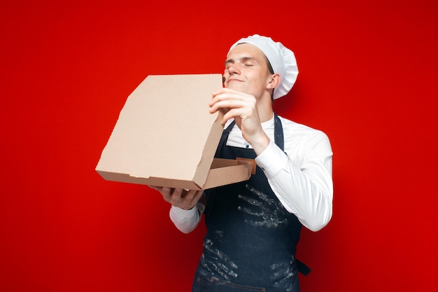 Baker holds pizza and sniffs on a red isolated background the chef with a box of pizza