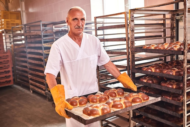 Baker houdt een dienblad met vers warm brood in zijn handen tegen de achtergrond van planken met gebak in de bakkerij Industriële productie van brood