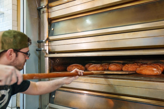 Foto baker's delight giovane uomo che mette abilmente le pagnotte di grano nel forno in un laboratorio di panetteria