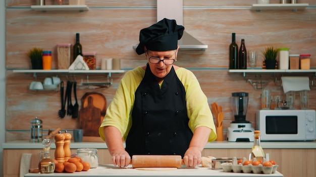 Baker woman rolling dough at home infront of video camera explaining recipe step by step. Retired blogger chef influencer using internet technology communicating on social media with digital equipment