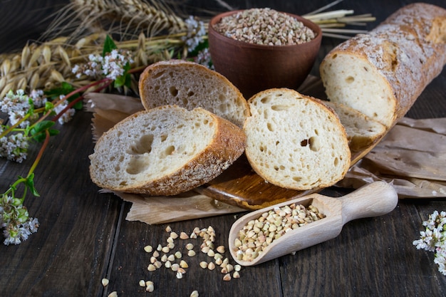 Bakery concept. Buckwheat bread, French baguette and stalks of wheat, oats, buckwheat on dark wooden table.