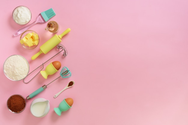 Bakery food frame, cooking concept. Ingredients on kitchen table. Butter, sugar, flour, eggs, oil, spoon, rolling pin, brush, whisk over pink background.