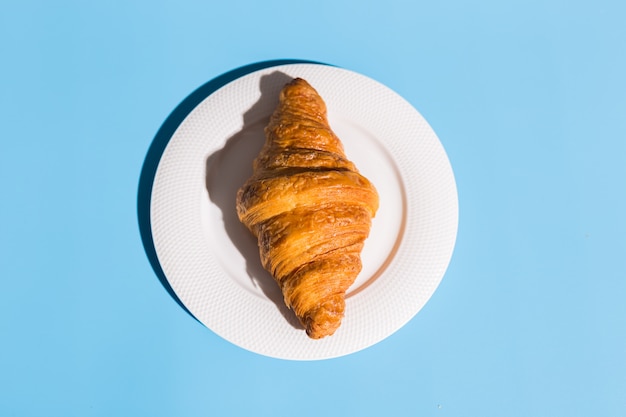 Photo bakery products baked croissant on white plate. blue background, top view close-up. pop art style. summer shadows. delicious and food concept.