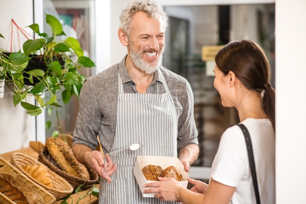 Bakery shop. Happy gray haired salesman showing packed buns to woman with long dark hair standing with back to camera