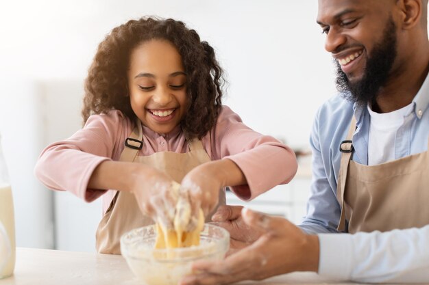 Baking. Closeup Portrait Of Excited Black Girl Kneading Dough With Hands In Kitchen Interior, Cheerful Dad Helping His Daughter, Family Having Fun While Preparing Homemade Pastry, Selective Focus
