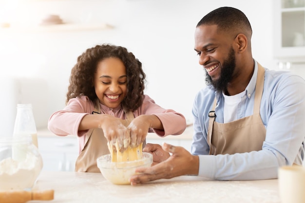 Concetto di cottura. ritratto di gioiosa ragazza nera che impasta la pasta con le mani nell'interiore della cucina, indossando il grembiule, papà allegro che aiuta sua figlia, famiglia divertendosi mentre prepara la pasticceria fatta in casa